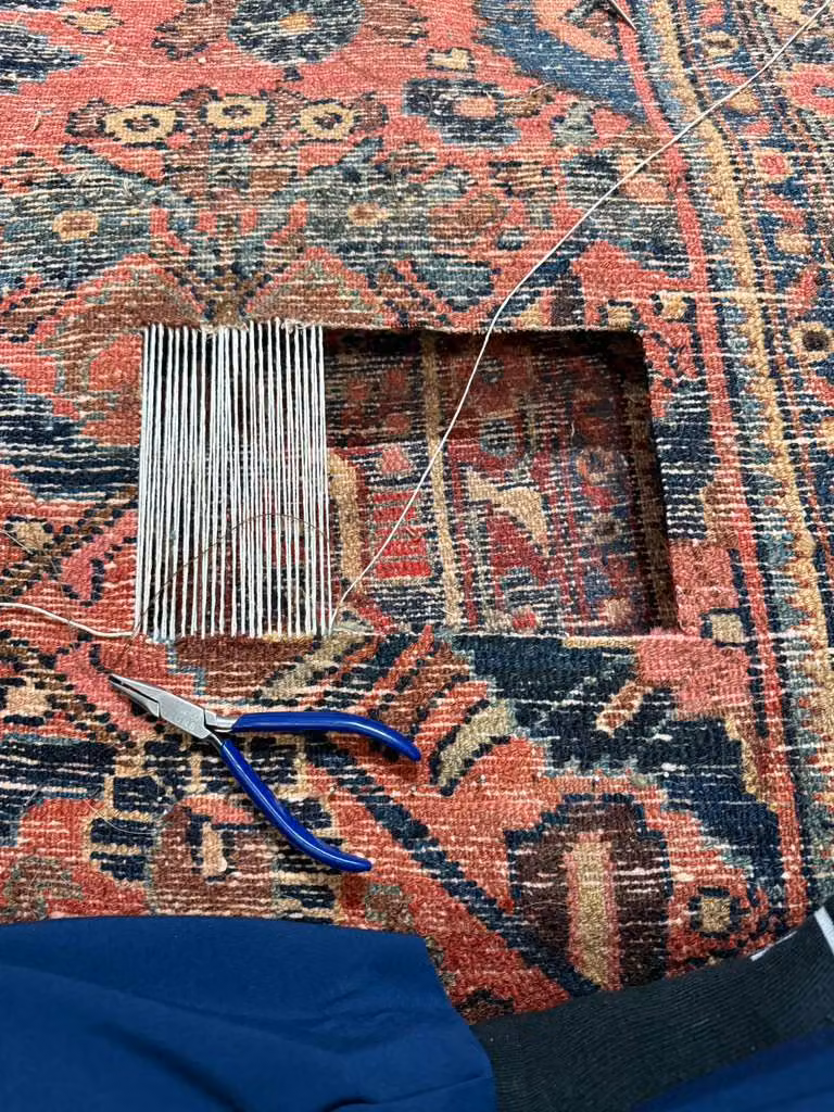 Close-up of a patterned rug with a rectangular repair opening, loose threads, a fringe-like repair piece, pliers, and a hand.